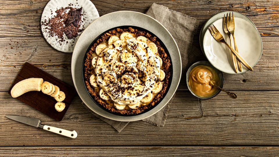 Selbstgemachter Butterkeks-Boden, aus Kondensmilch entstandenes Toffee, Bananenscheiben und eine Sahne-Crème-fraîche-Creme werden nur noch durch und mit Ahornsirup und Schokoladenraspeln getoppt.