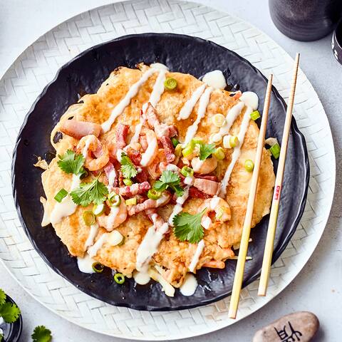 Okonomiyaki kommen aus Japan und sind eine Art dünner Pfannkuchen, der mit einer typischen Okonomiyaki-Sauce gewürzt und mit Speck, Zwiebeln und Garnelen belegt wird.