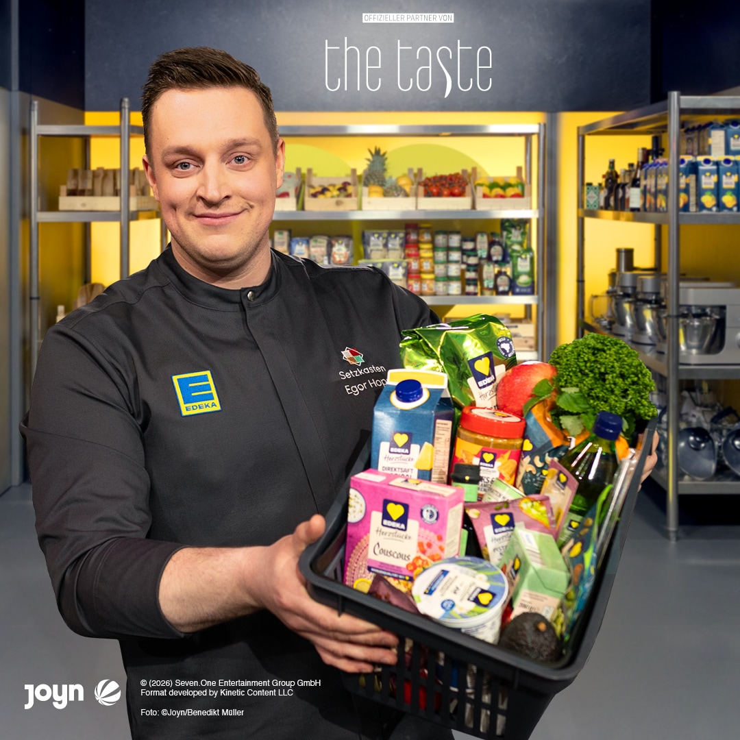Person in EDEKA-Kochjacke hält einen Korb mit EDEKA-Herzstücke-Produkten vor einer Speisekammer-Kulisse von „The Taste“.