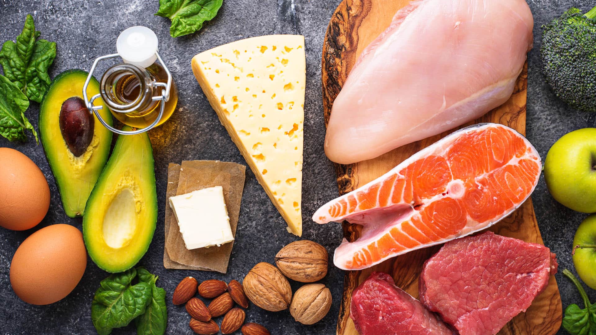 Avocado, Eier, Butter, Käse, Hähnchenbrust, Brokkoli, Apfel und Nüsse auf dunkler Unterlage