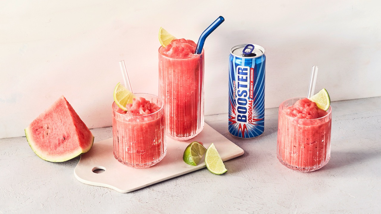 Drei mit rotem Slush-Ice gefüllte Gläser. Daneben ein Stück Wassermelone, zwei Limettenstücke sowie eine geöffnete Dose Booster Original.