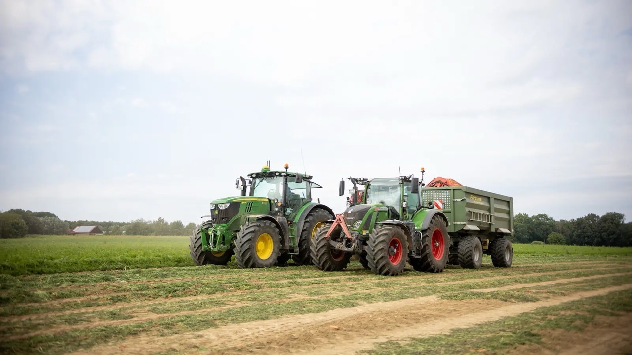 Zwei grüne Traktoren fahren über ein Feld. Einer zieht einen Anhänger.