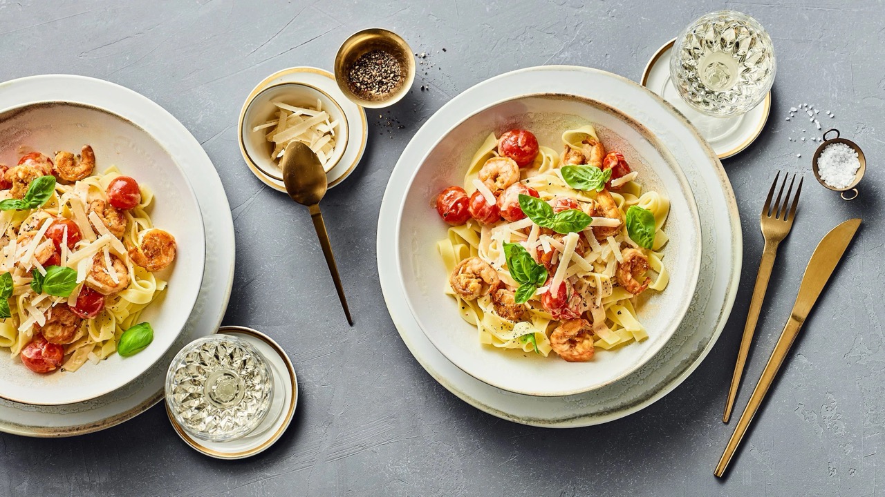 Zwei auf tiefen Tellern angerichtete Portionen Garnelen mit Tomaten auf Bandnudeln. Dazu Gläser, Besteck, grobes Salz, Pfeffer sowie Parmesan in Schälchen.