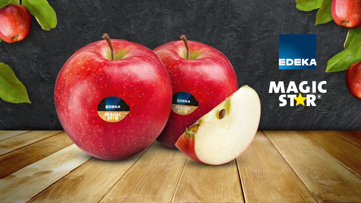 Zwei rote Äpfel der Sorte Magic Star mit EDEKA-Aufkleber und eine Apfelspalte auf einem Holztisch vor dunklem Steinuntergrund. Oben rechts das EDEKA-Logo und der Schriftzug „MAGIC STAR®“.