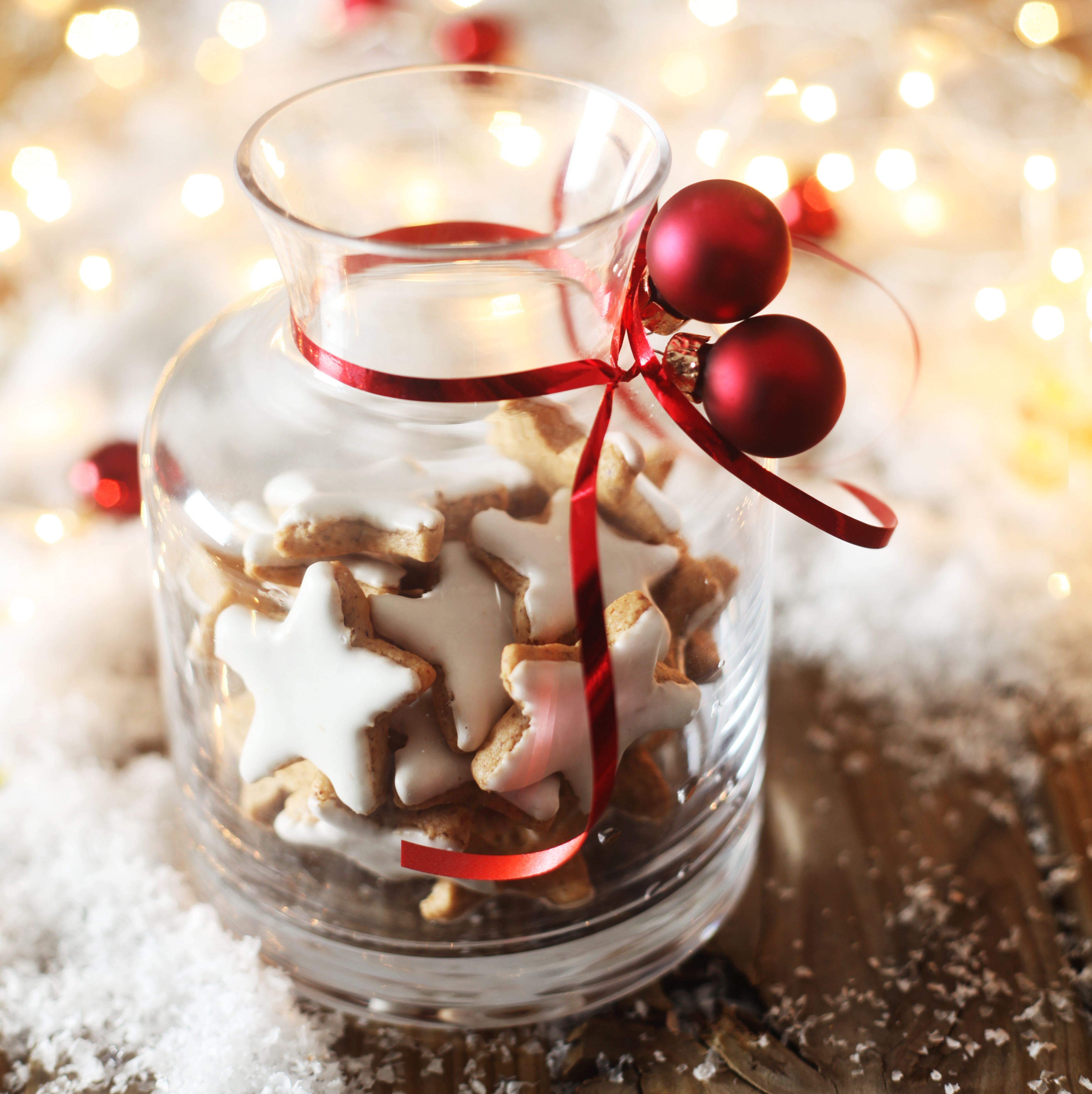 Festlich dekoriertes Glas mit selbst gebackenen, sternförmigen Weihnachtskeksen, verziert mit rotem Band und Weihnachtskugeln, auf einem Holztisch mit Kunstschnee und warmen Lichtern im Hintergrund.