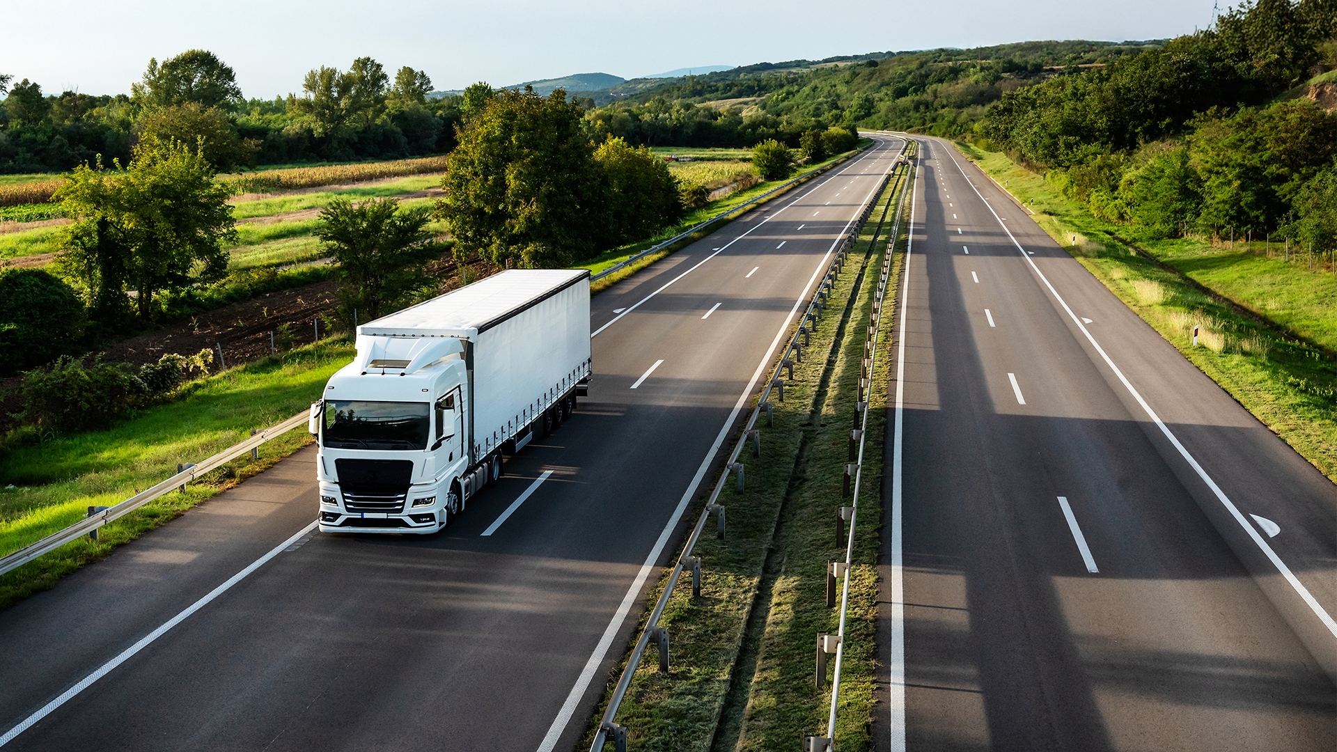 Ein LKW fährt auf einer leeren Straßen zwischen Wiesen und Bäumen, in hügeliger Landschaft.