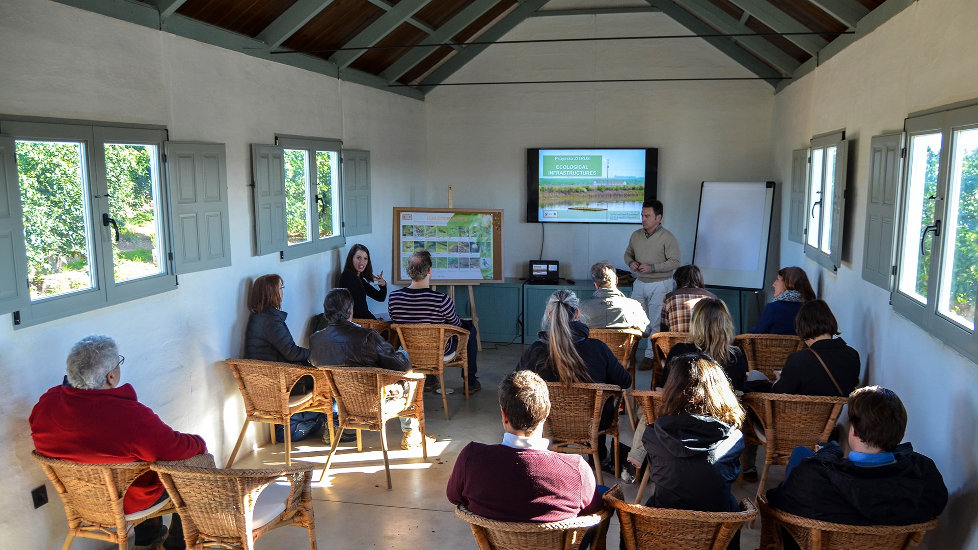 Eine Gruppe von Menschen, die in einem Raum sitzen und frontal einer Person zuhören