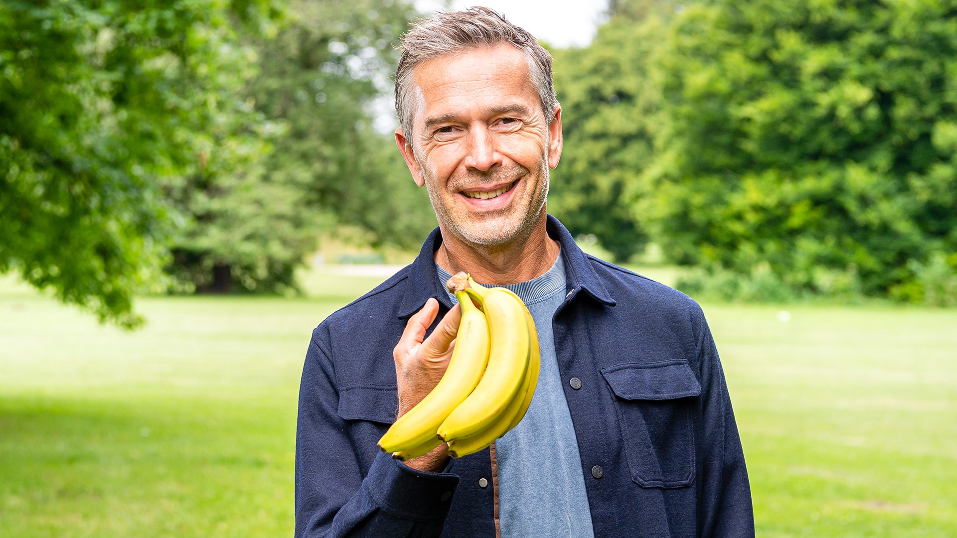 Dirk Steffens hält eine Bananestaude auf einer grünen Wiese in der Hand