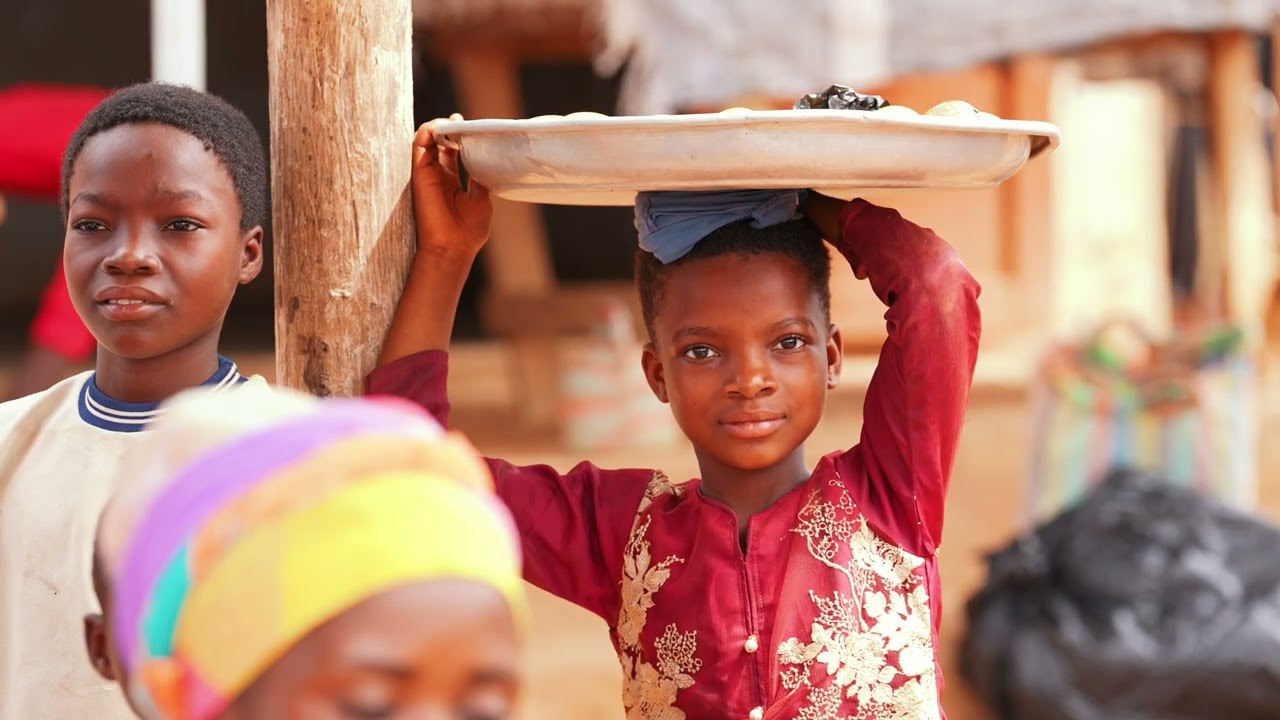Ein Kind trägt ein Tablett auf dem Kopf, weitere Kinder sind im Bild sichtbar