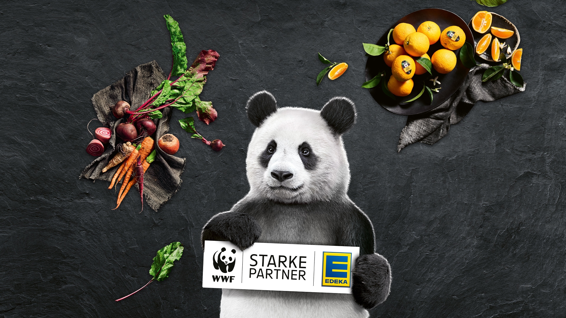Panda hält Schild mit den WWF und EDEKA Logos und "Starke Partner", umgeben von frischem Bio-Gemüse wie Roter Bete, Karotten und Mangold auf einem braunen Leinentuch sowie Orangen in einer schwarzen Schale.