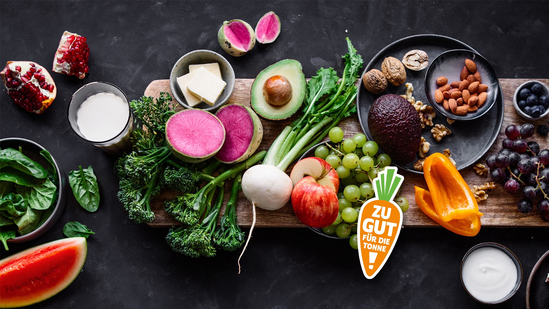 Verschiedene Obst-, Gemüse- und Milchprodukte auf einem Tisch, darunter Avocado, Brokkoli, Trauben, Paprika und Nüsse, mit dem Hinweis „Zu gut für die Tonne!“.