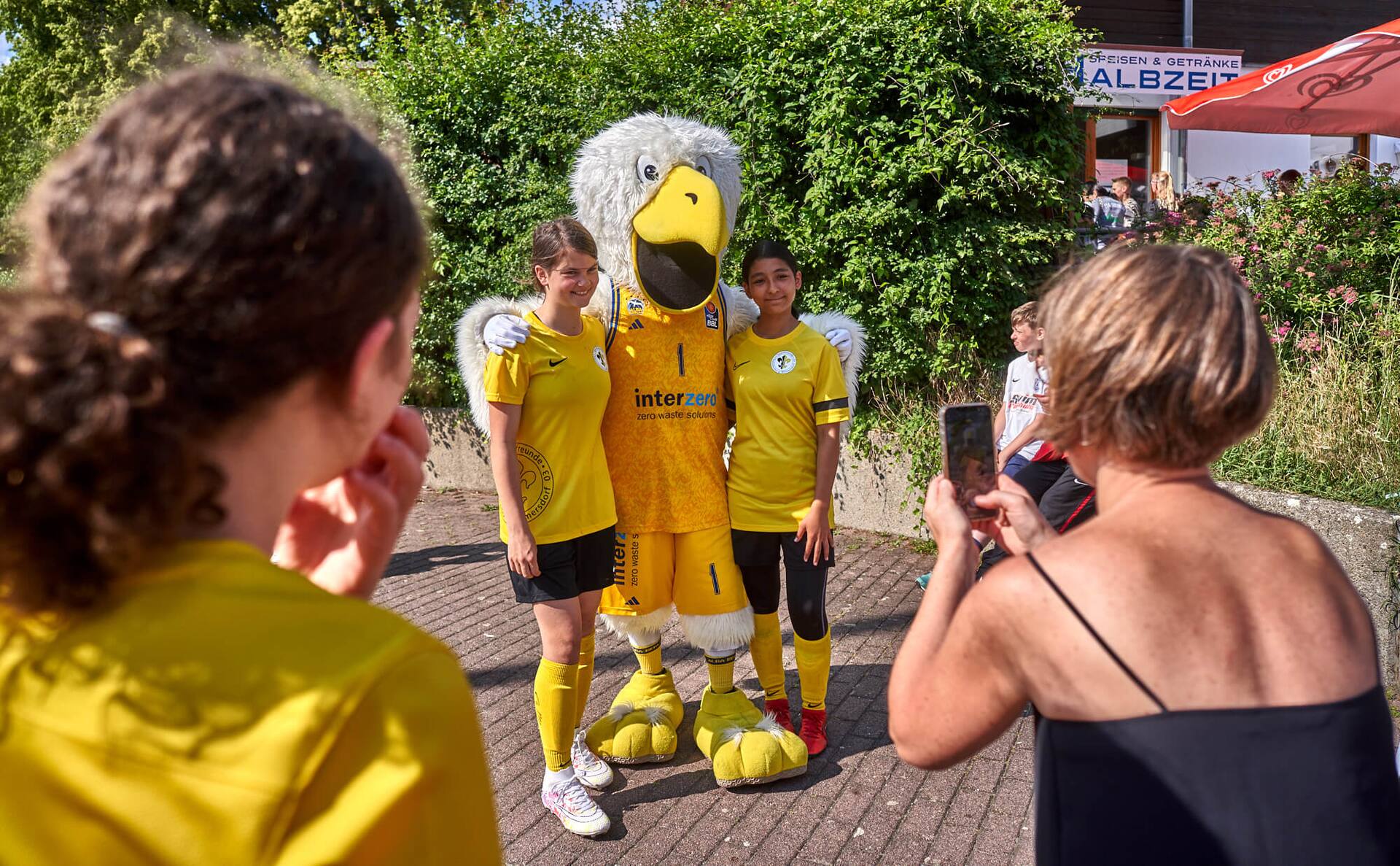 Albatros-Maskottchen mit gelber Sportkleidung zwischen zwei Jugend-Fußballspielerinnen.