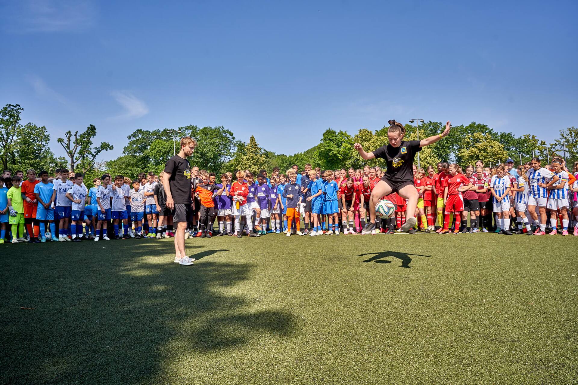 Eine Fußball-Spielerin macht Tricks mit einem Ball. Hinter ihr stehen verschiedene Mannschaften und schauen ihr zu.