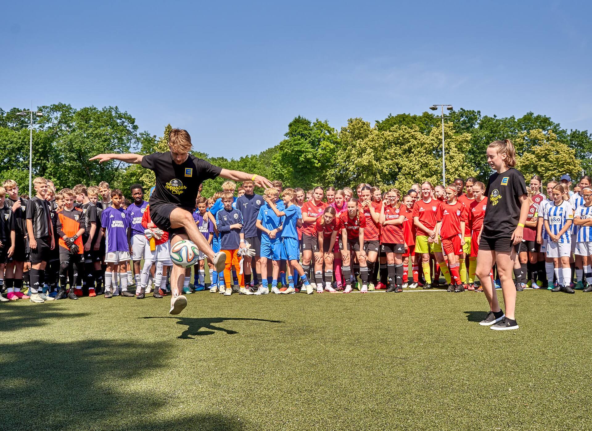 Eine Fußball-Spieler macht Tricks mit einem Ball. Hinter ihm stehen verschiedene Mannschaften und schauen ihr zu.