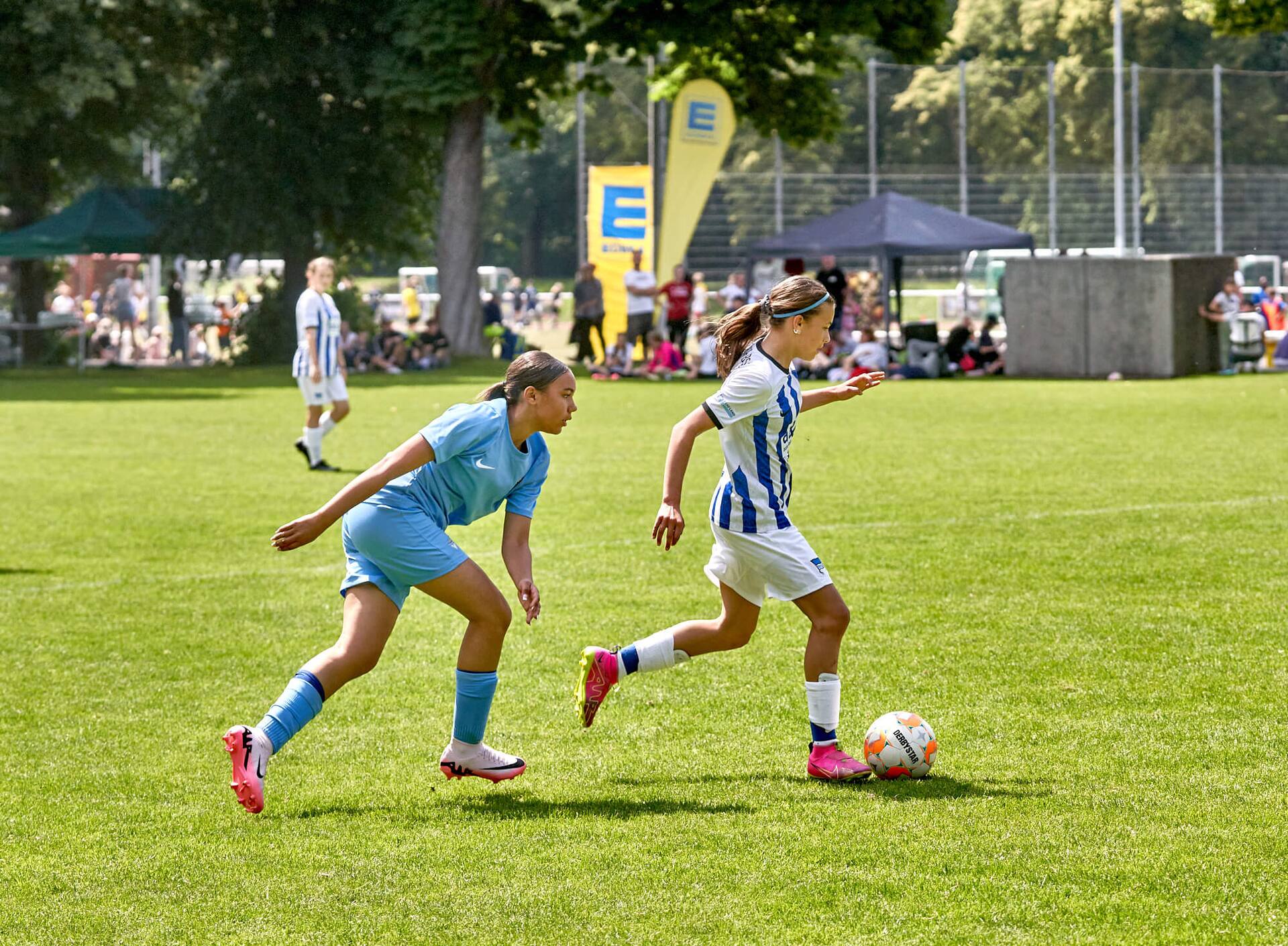 Eine Jugend-Fußballspielerin in blau-weiß gestreiftem Trikot läuft mit dem Ball am Fuß. Die Gegenspielerin hinter ihr ist ins Straucheln geraten.