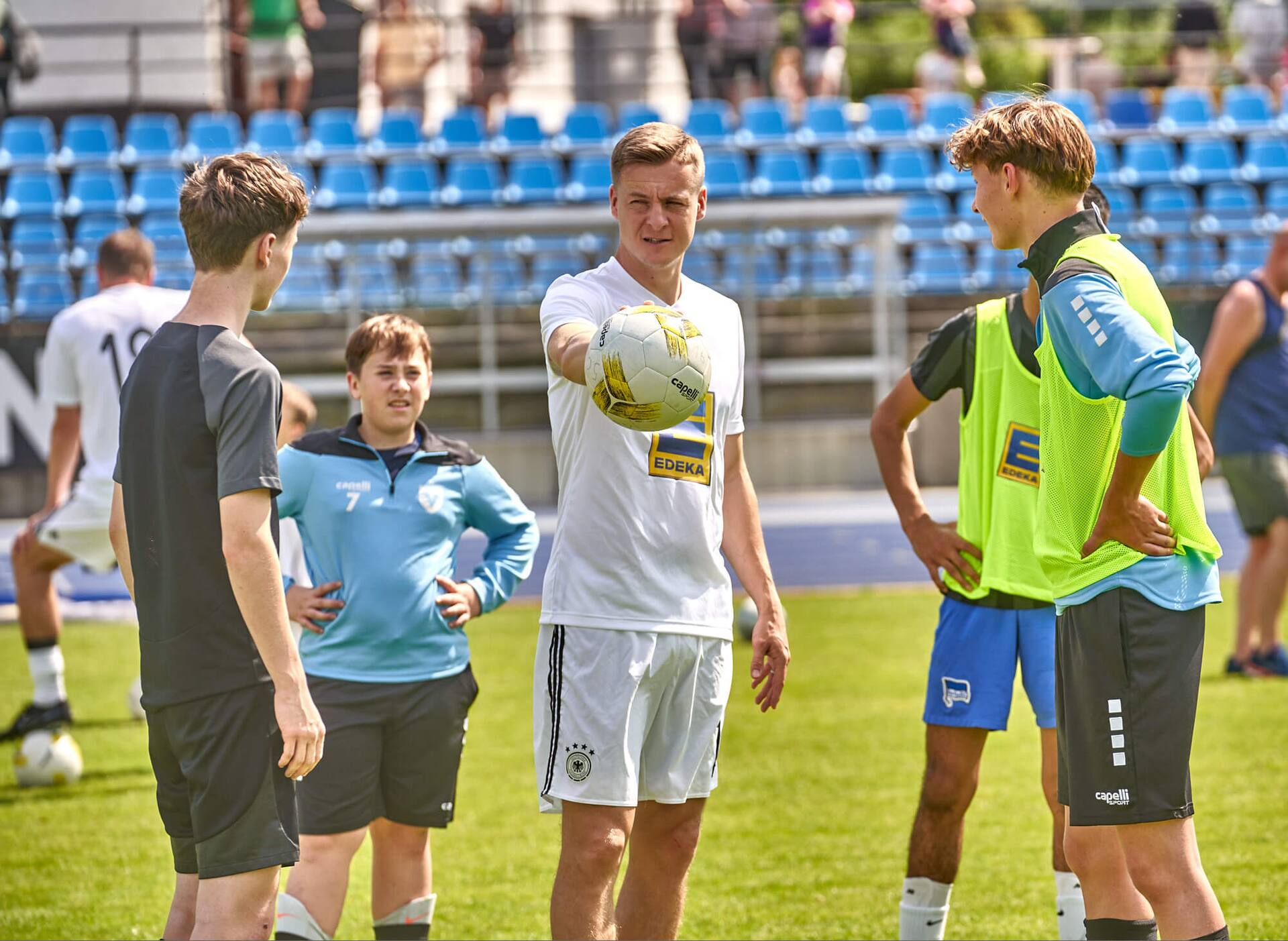 Ex-Profi Felix Kroos hält einen Ball in der Hand. Neben ihm stehen zwei männliche Jugend-Fußballer