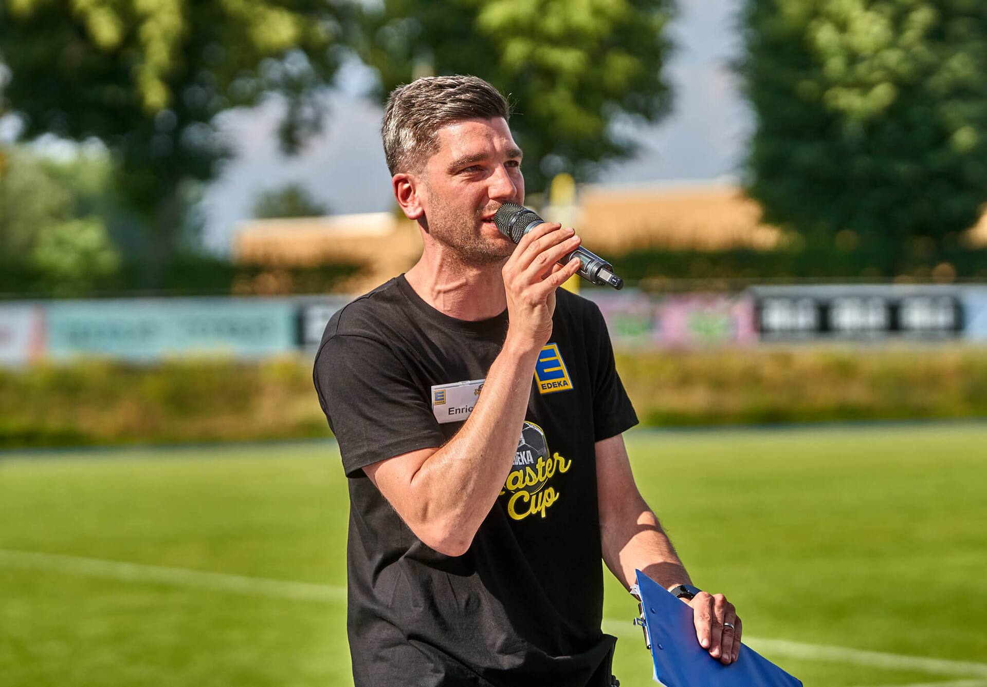Mann mit Mikrofon in der Hand. Er trägt ein schwarzes T-Shirt mit Aufdruck EDEKA Master Cup.