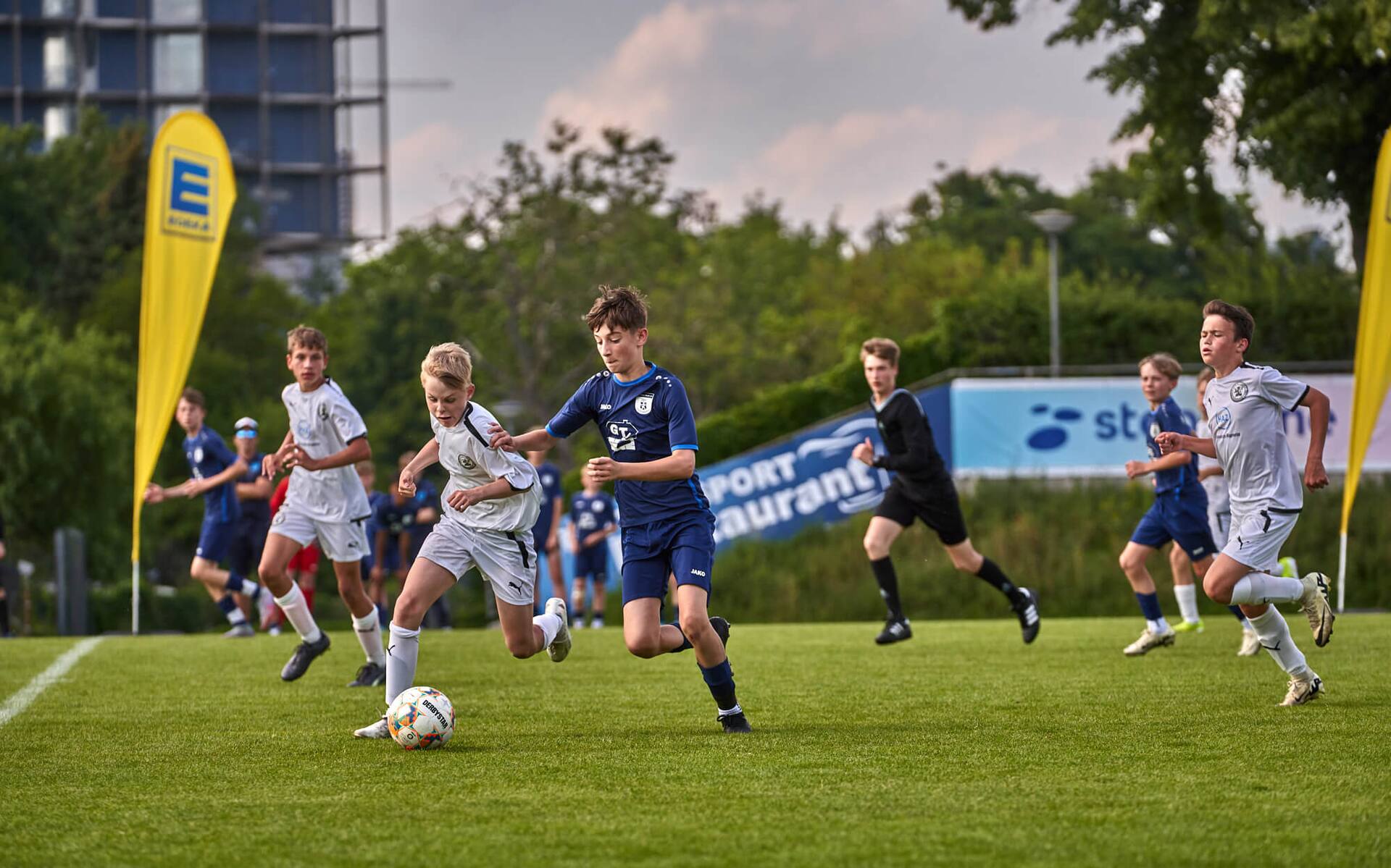 Dynamischer Zweikampf um den Ball zwischen zwei Jugendspielern in weißen und blauen Trikots.
