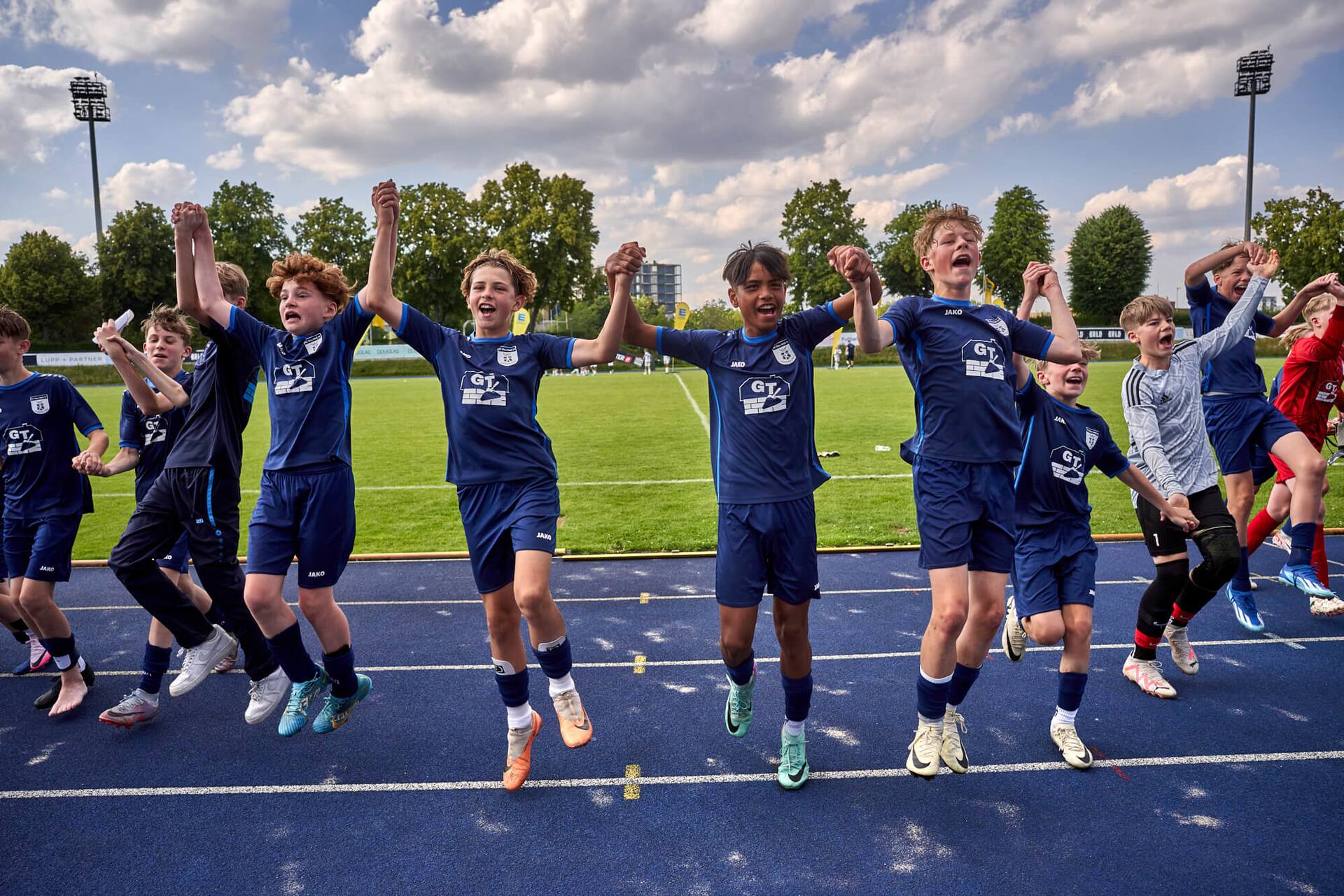 Männliches Jugend-Fußballteam hält sich in den Händen und mach Jubelsprünge auf einer blauen Laufbahn.