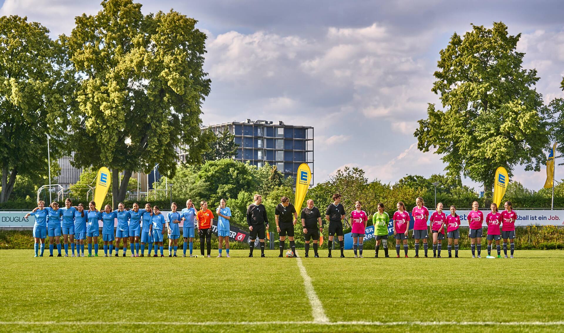 Zwei weibliche Jugend-Fußballteams haben sich vor dem Spiel am Mittelkreis aufgestellt. Zwischen den Teams stehen vier Schiedsrichter.