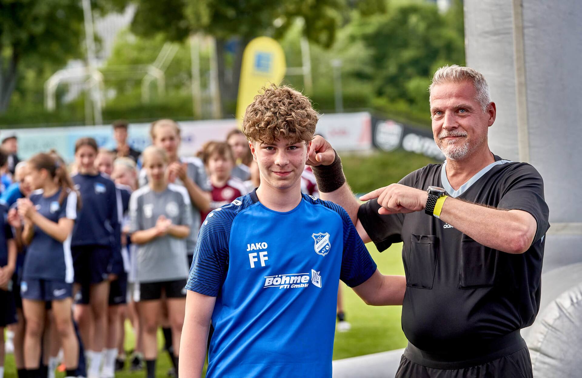 Ein Schiedsrichter zeigt außerhalb des Spielfeldes auf einen männlichen Jugend-Fußballspieler in einem blauen Trikot.