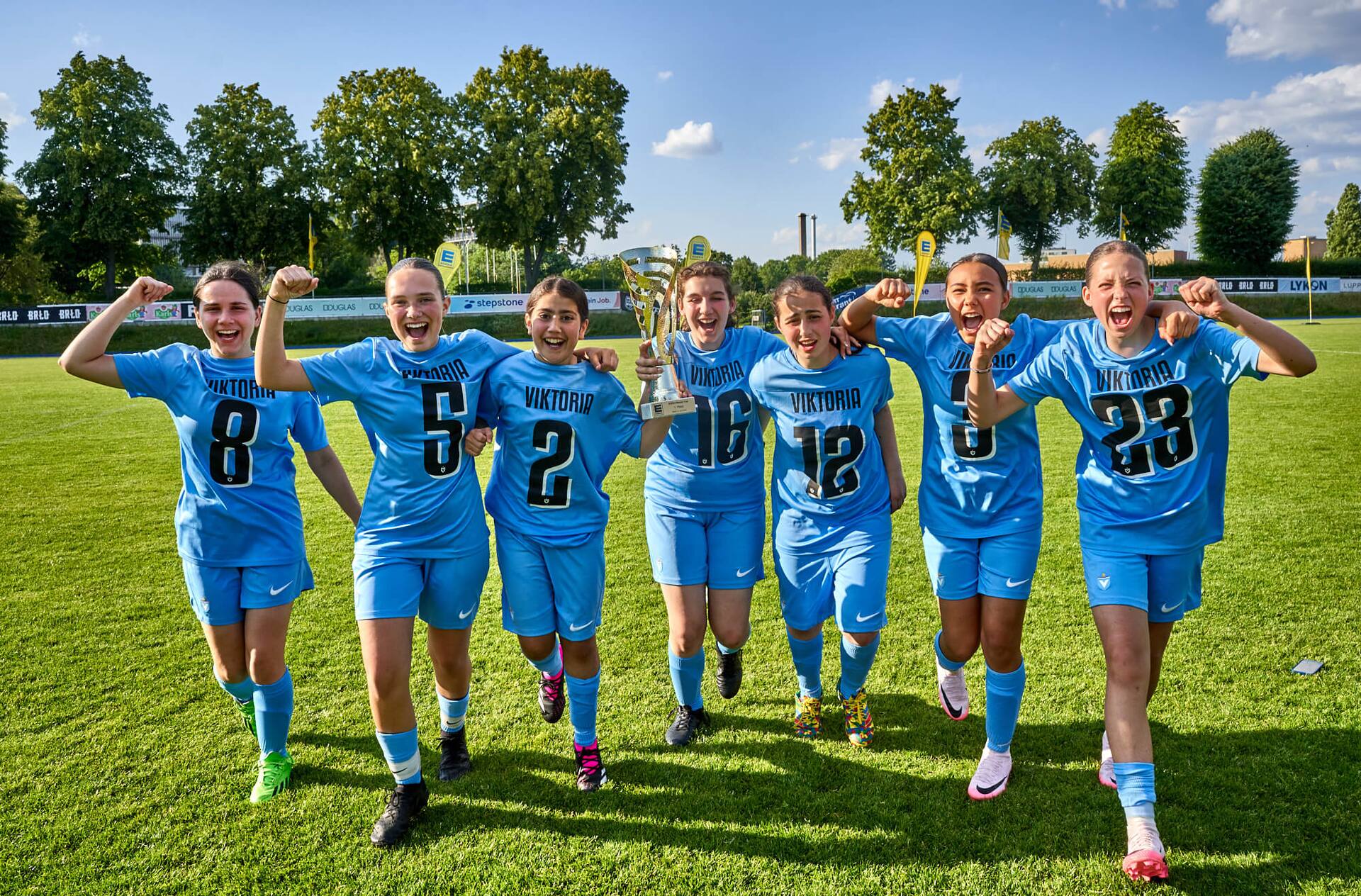 Sieben Spielerinnen von Viktoria laufen jubelnd mit einem Pokal in der Hand.