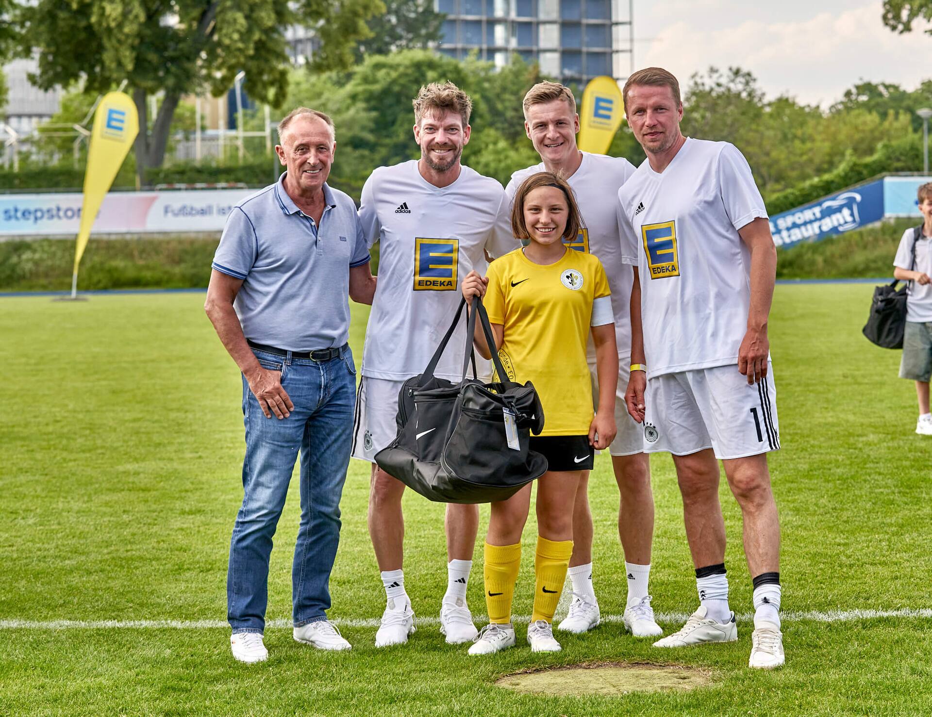 Drei Ex-Profis und ein älterer Herr stehen mit einer Jugendfußballspielerin zusammen. Sie hält eine große neue Sporttasche in der Hand.
