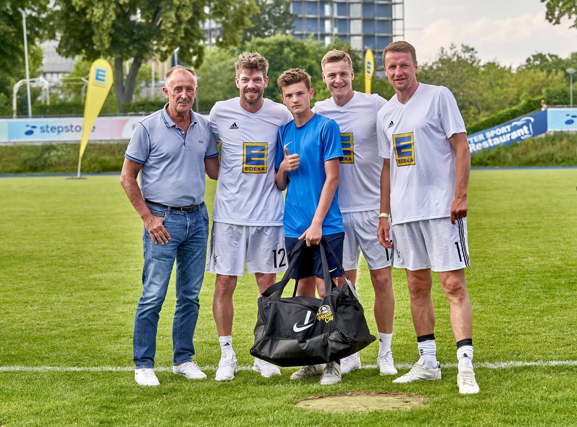 Drei Ex-Profis und ein älterer Herr stehen mit einem Jugendfußballspieler zusammen. Er hält eine große neue Sporttasche in der Hand.