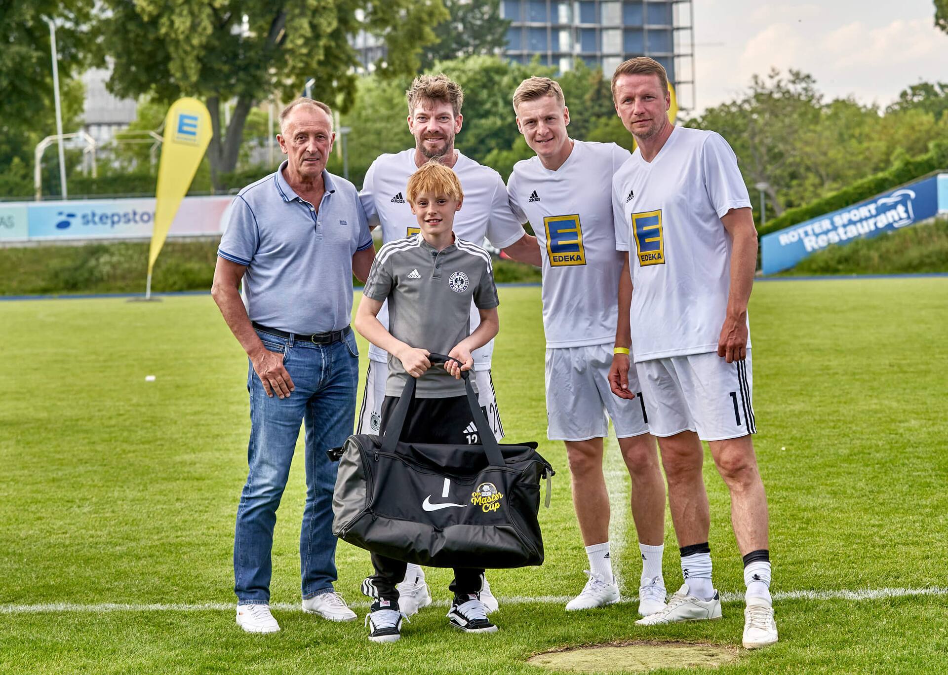 Drei Ex-Profis und ein älterer Herr stehen mit einem Jugendfußballspieler zusammen. Er hält eine große neue Sporttasche in der Hand.