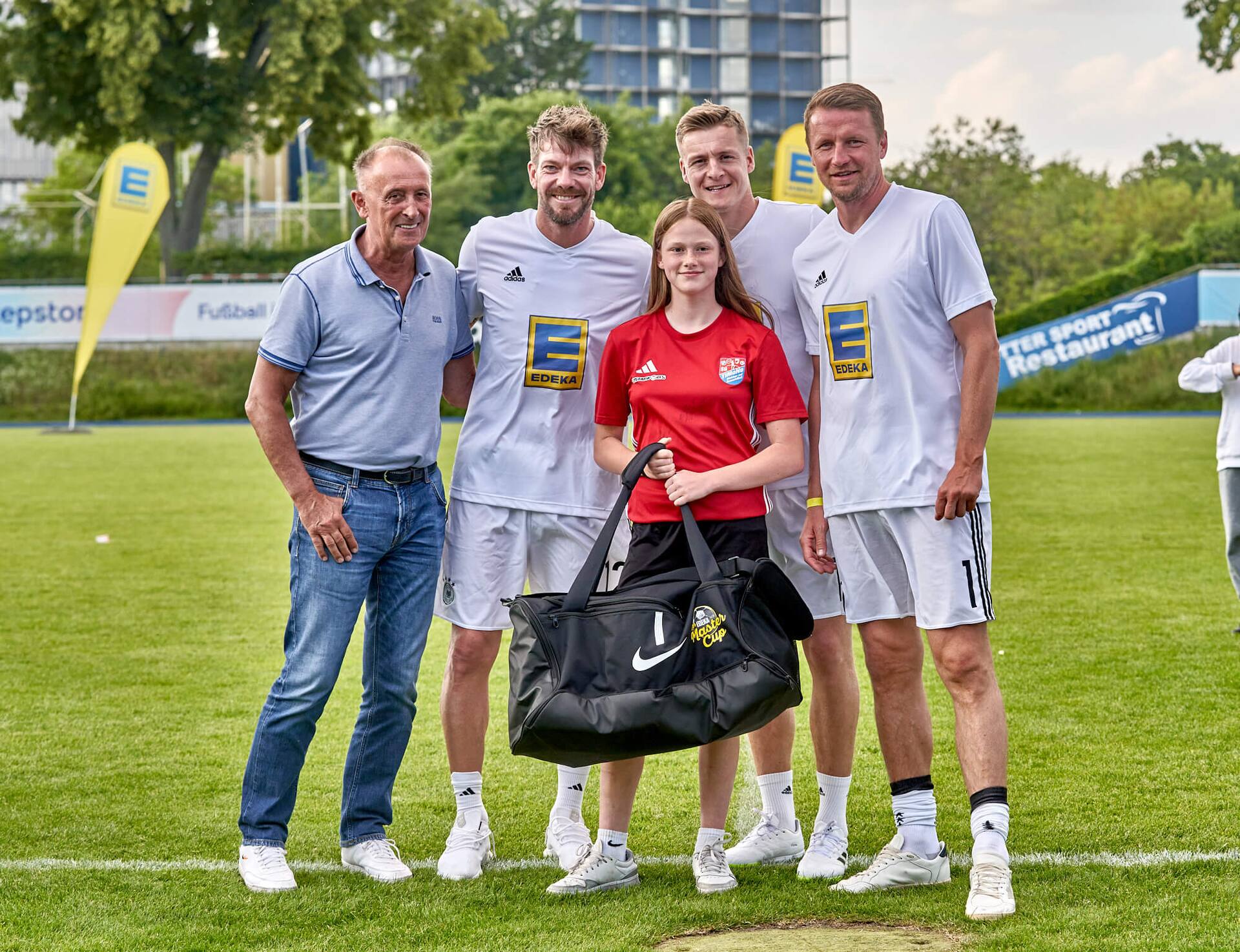 Drei Ex-Profis und ein älterer Herr stehen mit einer Jugendfußballspielerin zusammen. Sie hält eine große neue Sporttasche in der Hand.