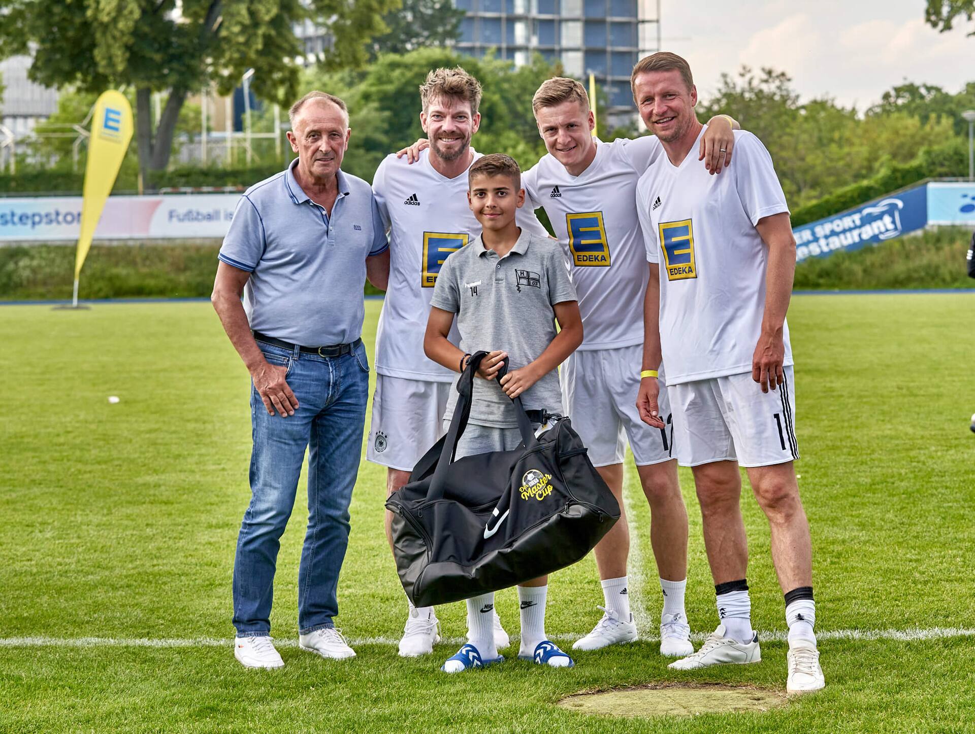 Drei Ex-Profis und ein älterer Herr stehen mit einem Jugendfußballspieler zusammen. Er hält eine große neue Sporttasche in der Hand.