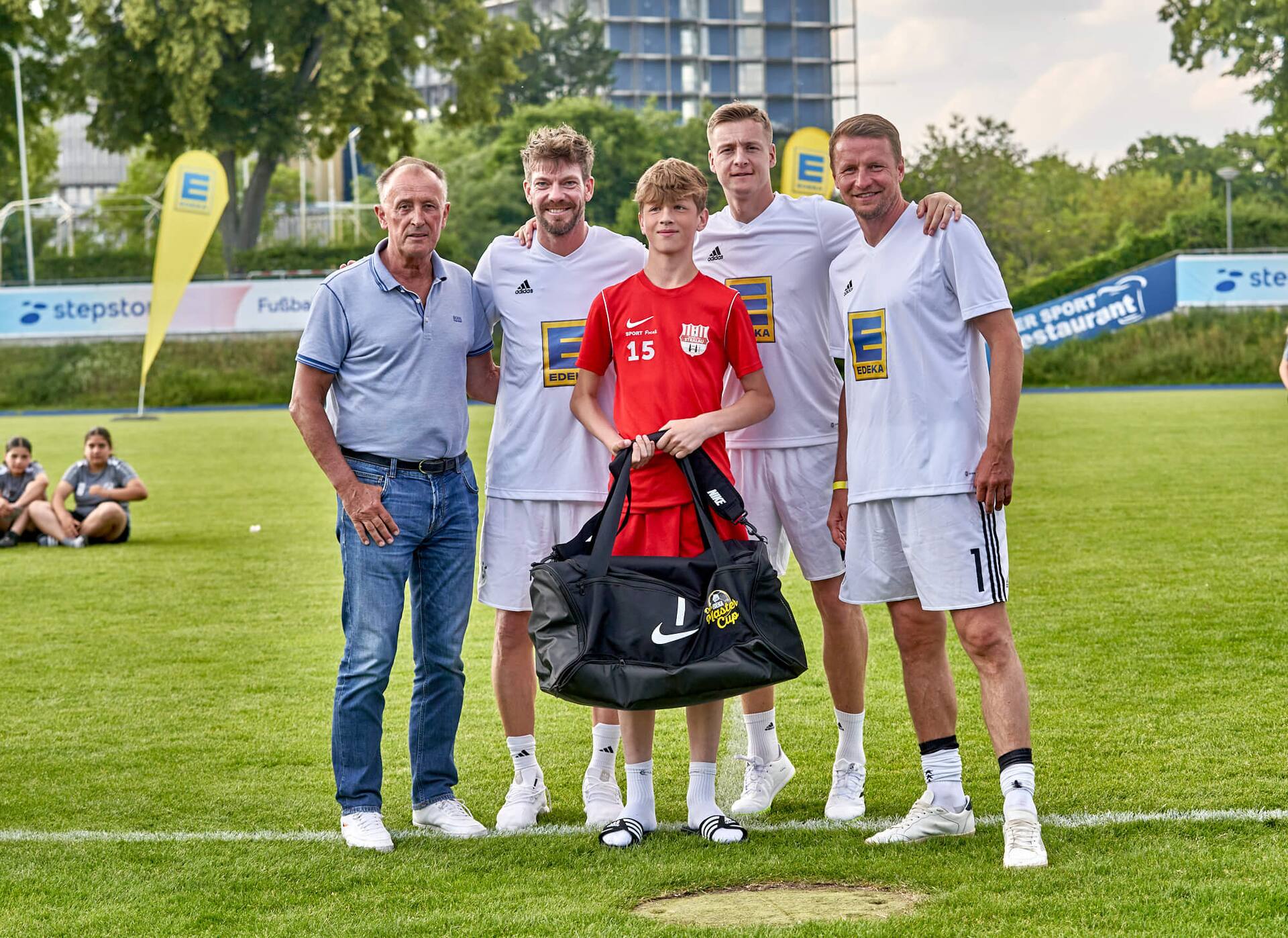 Drei Ex-Profis und ein älterer Herr stehen mit einem Jugendfußballspieler zusammen. Er hält eine große neue Sporttasche in der Hand.
