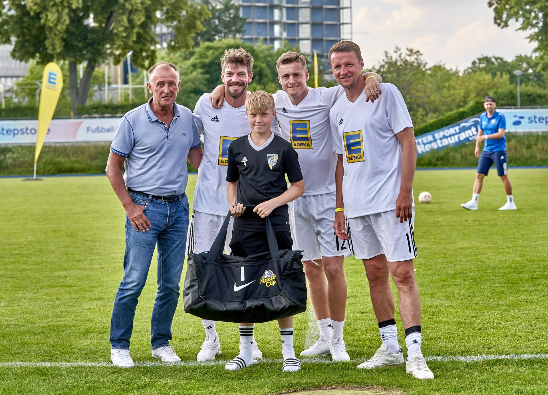 Drei Ex-Profis und ein älterer Herr stehen mit einem Jugendfußballspieler zusammen. Er hält eine große neue Sporttasche in der Hand.