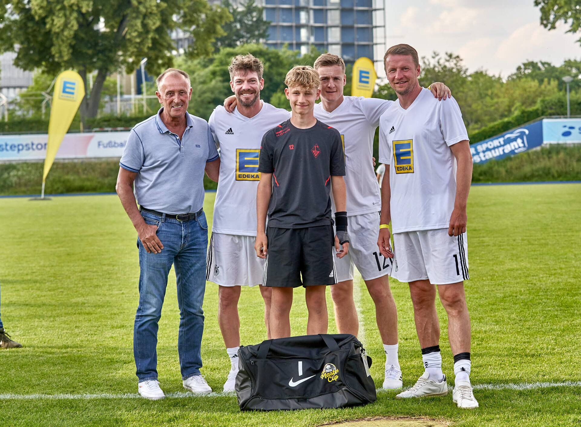 Drei Ex-Profis und ein älterer Herr stehen mit einem Jugendfußballspieler zusammen. Vor ihm steht eine große neue Sporttasche.