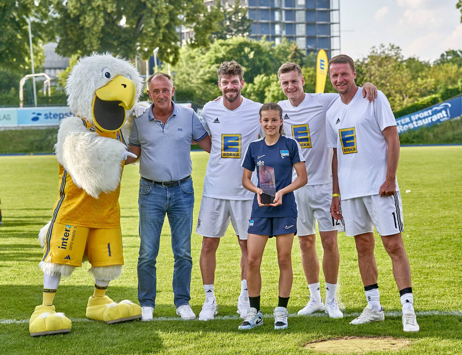 Ein Albatros-Maskottchen, drei Ex-Profis und ein älterer Herr stehen mit einer Jugendfußballspielerin zusammen. Sie hält einen Pokal in der Hand, auf dem „beste Spielerin“ steht.