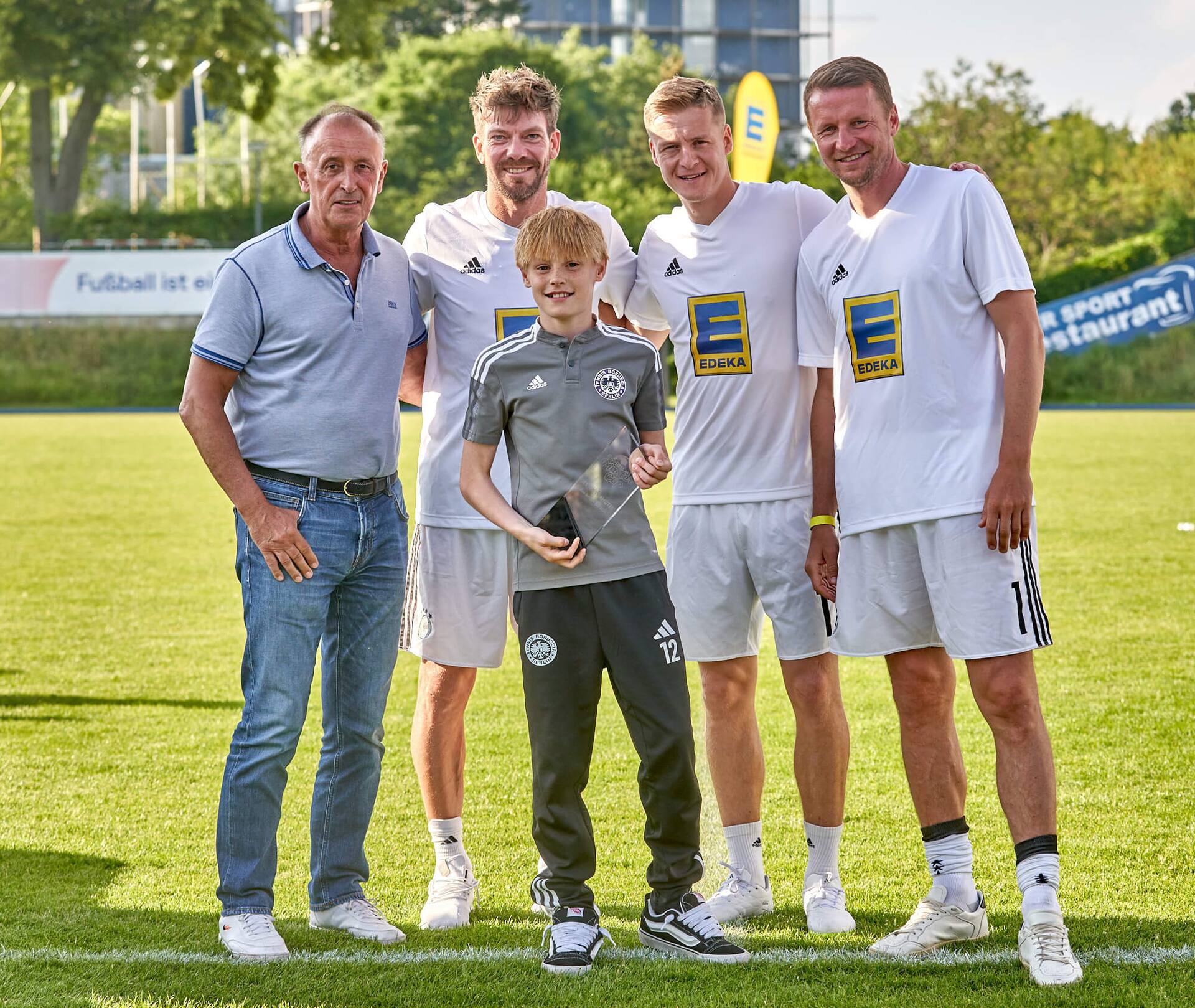 Ein Albatros-Maskottchen, drei Ex-Profis und ein älterer Herr stehen mit einem Jugendfußballspieler zusammen. Er hält einen Pokal in der Hand.