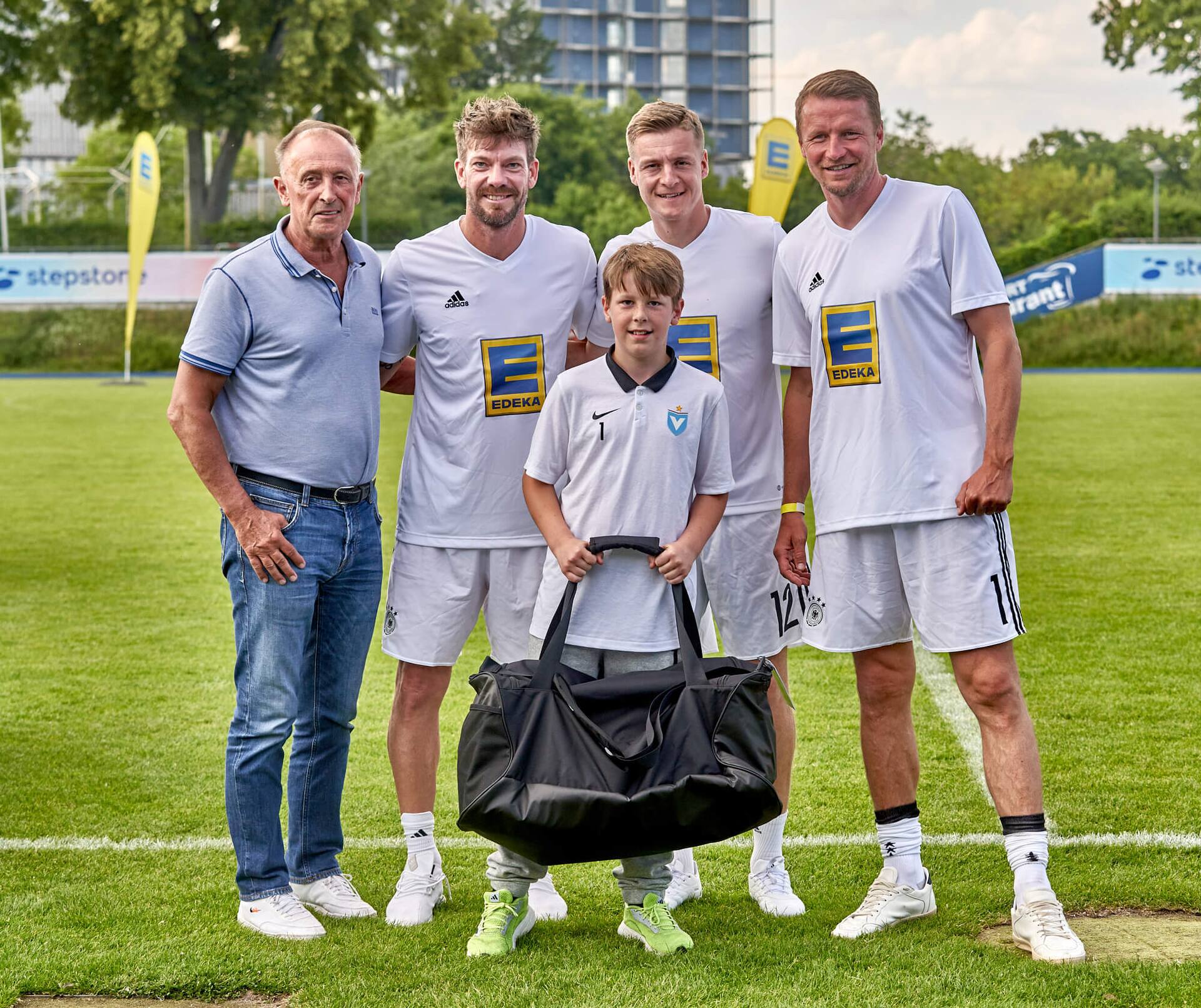 Drei Ex-Profis und ein älterer Herr stehen mit einem Jugendfußballspieler zusammen. Er hält eine große neue Sporttasche in der Hand.