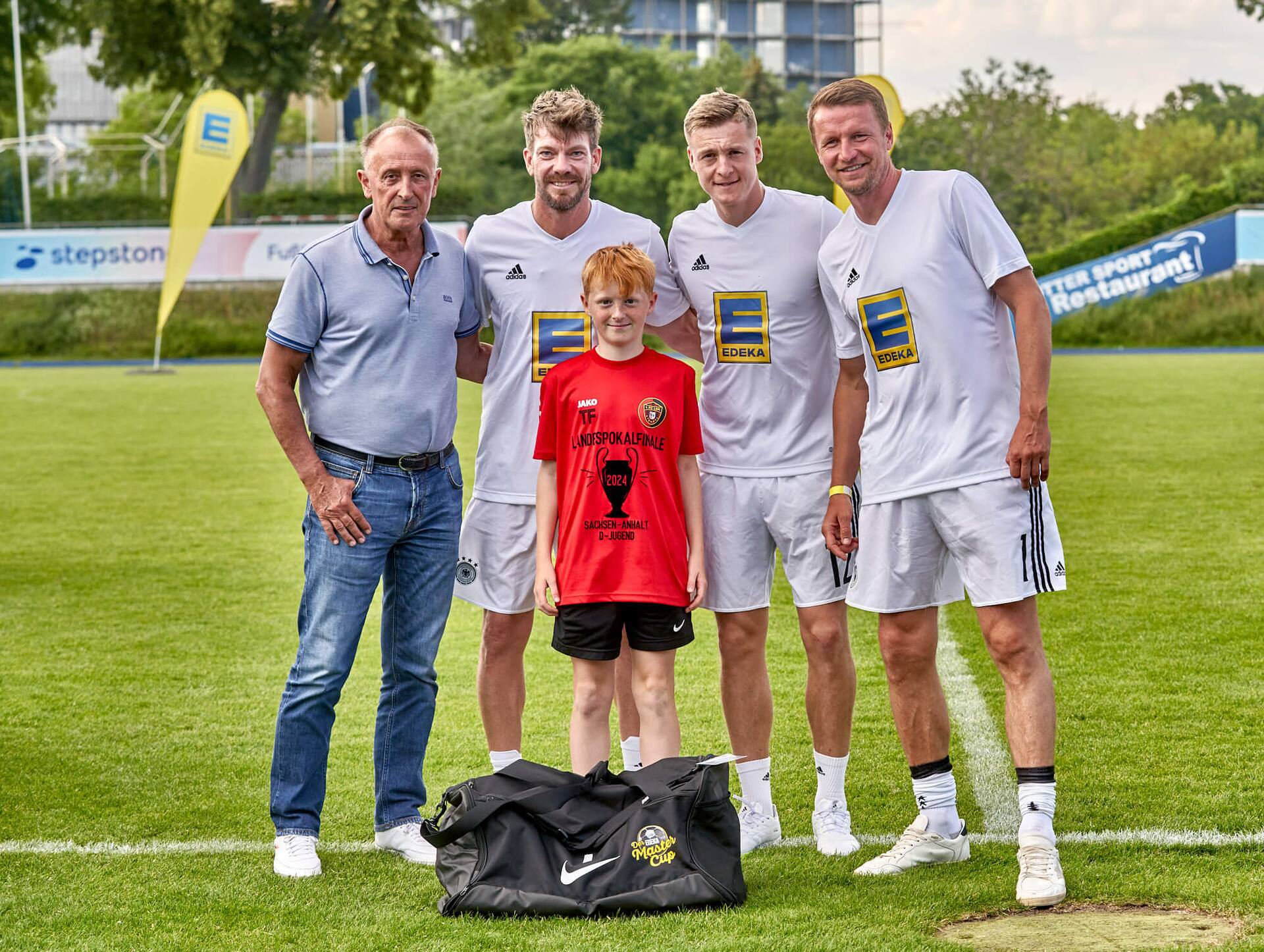 Drei Ex-Profis und ein älterer Herr stehen mit einem Jugendfußballspieler zusammen. Vor ihm steht eine große neue Sporttasche auf dem Rasen.