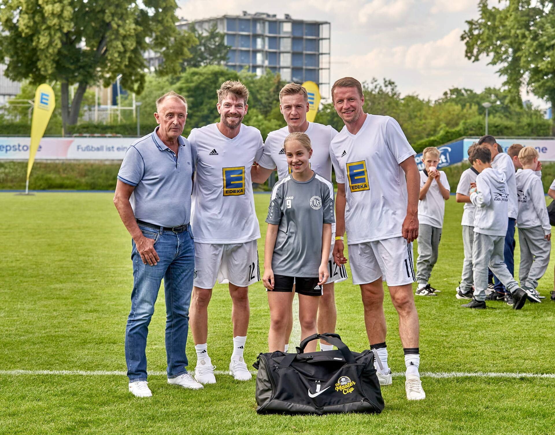 Drei Ex-Profis und ein älterer Herr stehen mit einer Jugendfußballspielerin zusammen. Vor ihr steht eine große neue Sporttasche auf dem Rasen.