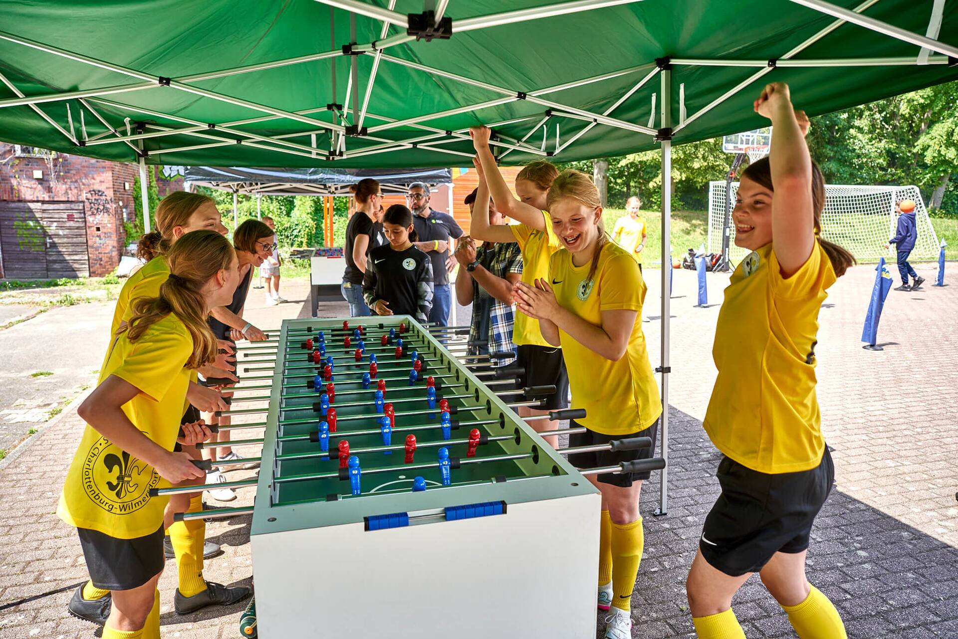Acht Personen spielen ausgelassen an einem überlangen Tischkicker unter einem grünen Pavillon.