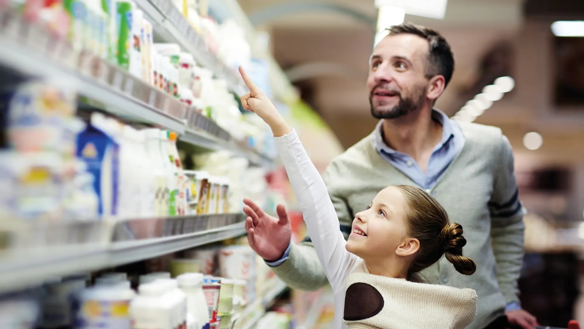 Das Bild zeigt einen Vater mit seinem Kind, seiner Tochter, die vor einem Kühlregal im Supermarkt stehen und die Tochter zeigt auf ein Produkt oben in dem Regal, oberhalb ihres Kopfes.