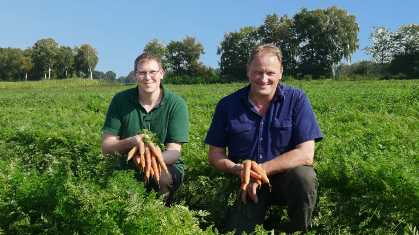 Zwei Männer hocken in einem Möhrenfeld. Sie lächeln und halten Bundmöhren in ihren Händen.