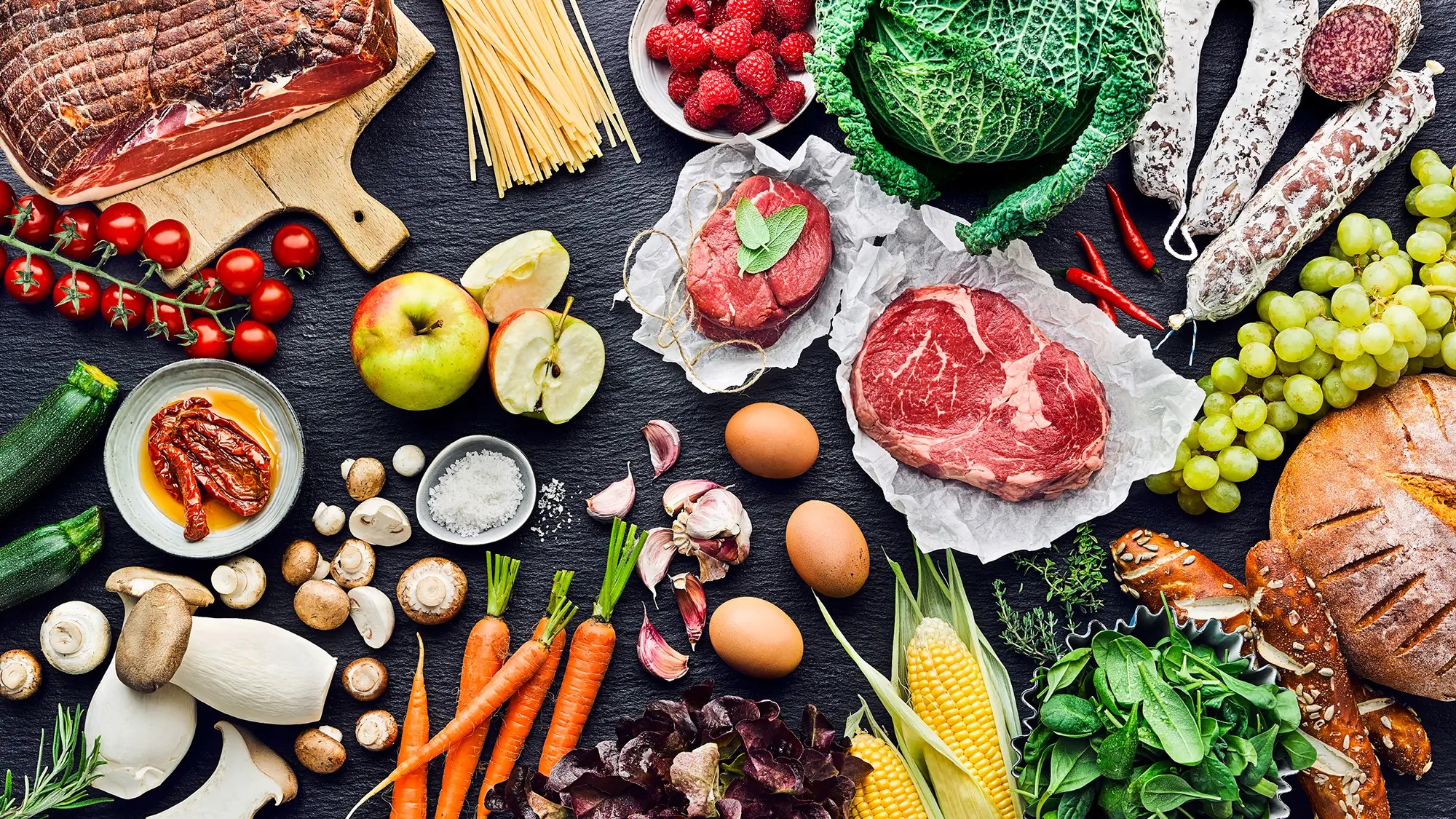 EDEKA Schiefertafel mmit verschiedenen Lenbensmitteln wie Fleisch & Wusr, Obst & Gemüse, Backwaren & Nudeln, Eier, eingelegte getrocknete Tomaten. Teilweise isnd die Lebensmittel angeschnitten, liegen in Schalen oder auf Brettern angerichtet auf der Platt oder sind noch ganz.