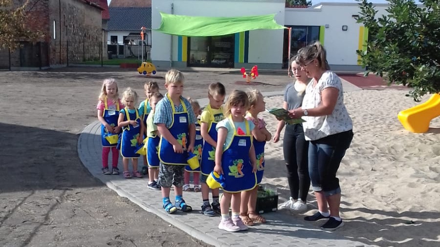 Das Bild zeigt Kinder in verschiedenen Altersstufen mit Schürzen und Gießkannen. Daneben stehen zwei Erzieherinnen und singen.