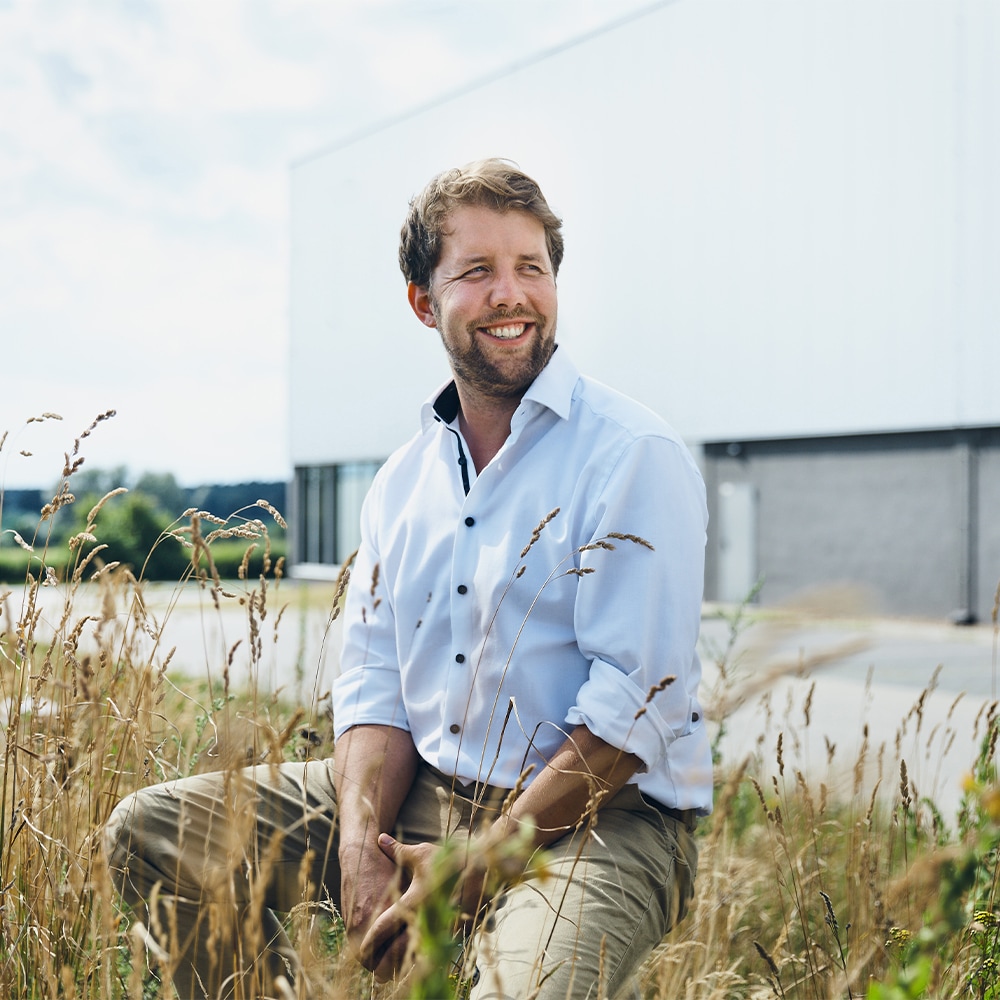 Zwischen Getreidehalmen sitzt ein lächelnder Mann in hellblauem vor einem großen weißen Gebäude.
