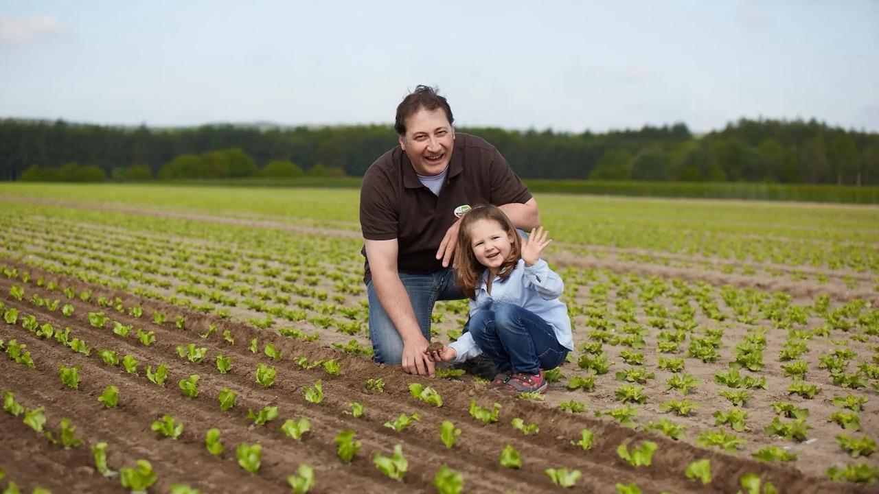 Ein Mann hock in einem Salatfeld und lacht. Neben ihm hockt ein winkendes Mädchen mit Erde in der Hand.