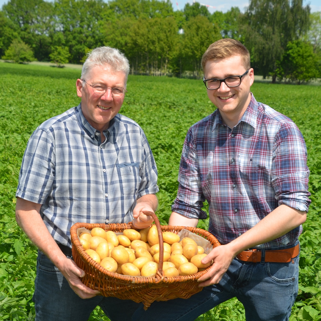 Ein jüngerer und ein älterer Herr stehen in einem Feld mit grünen Pflanzen. Sie halten einen Korb mit Kartoffeln.