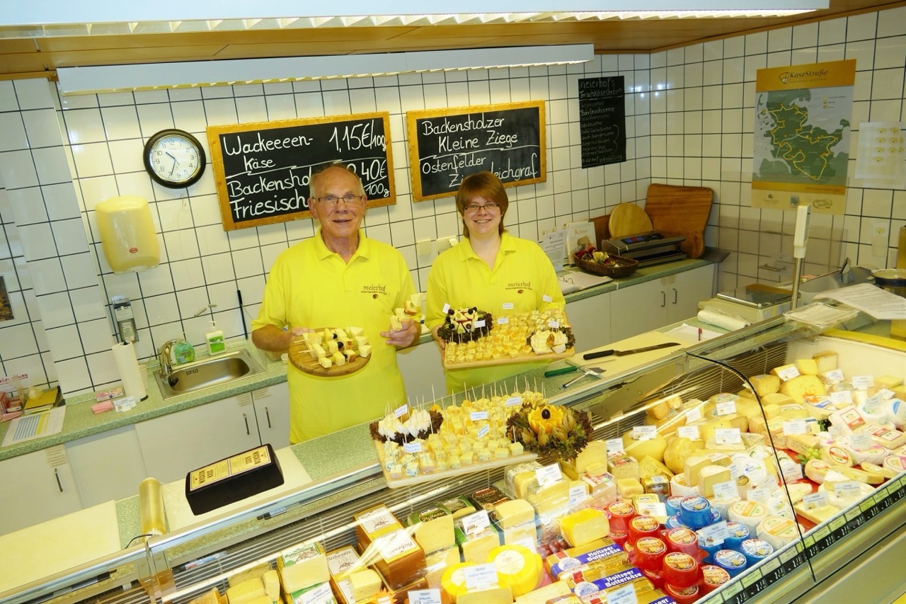 Zwei Personen in gelben Poloshirts stehen hinter einer Käsetheke. In ihren Händen halten sie Platten mit verschiedenen Käsespießen.