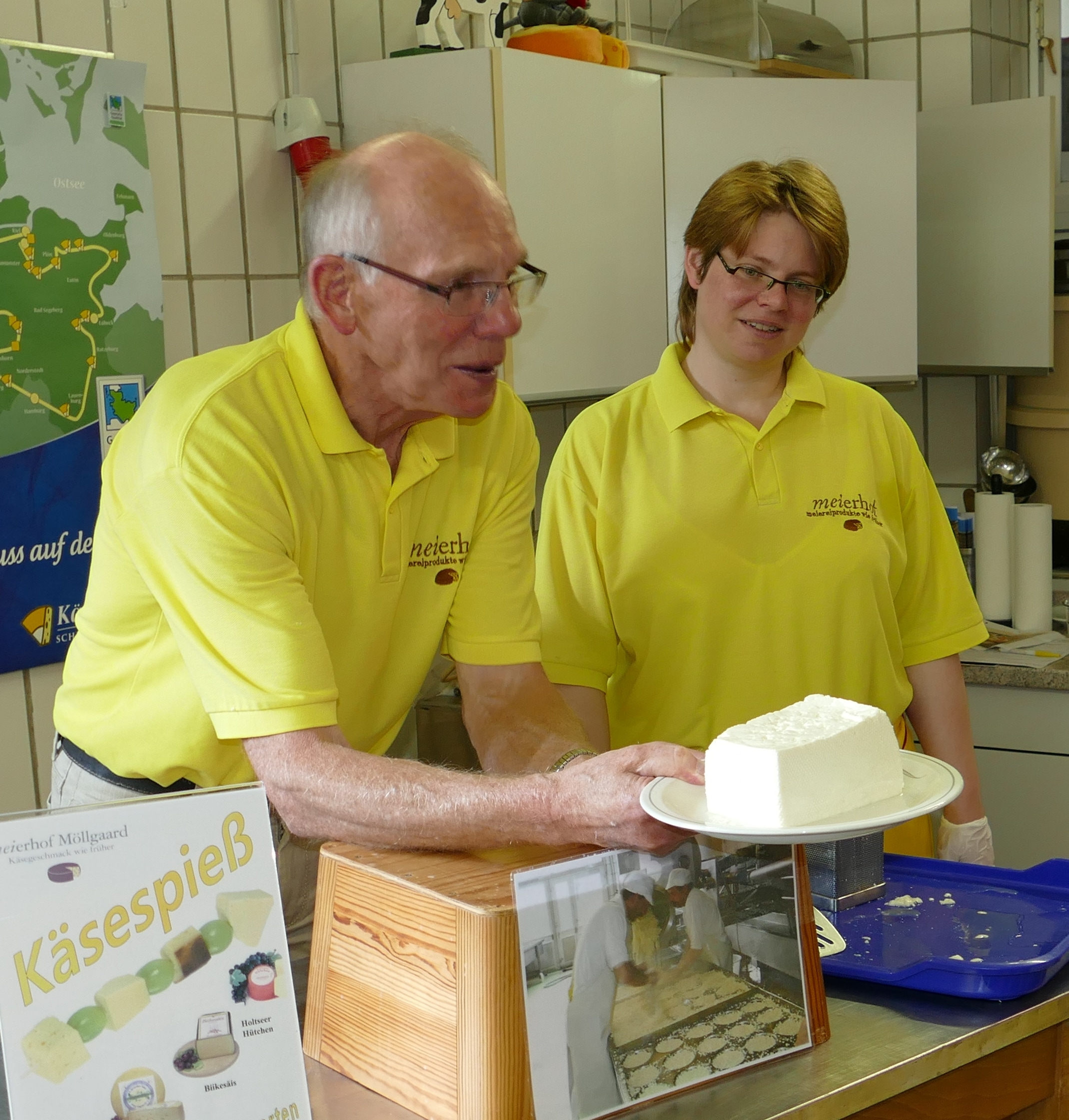 Zwei Personen in gelben Poloshirts stehen hinter einer Käsetheke. Der Herr links hält einen Teller mit einem großen weißen Stück Käse.