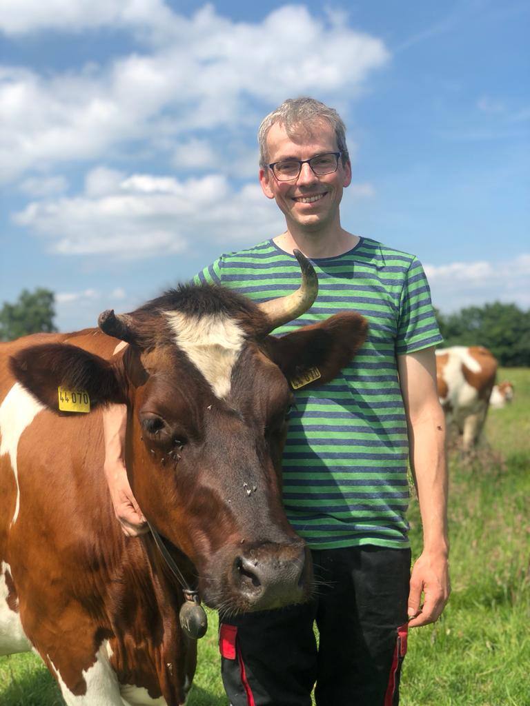 Ein lächelnder Mann mit Brille im grün-blau gestreiften T-Shirt steht neben einer braunen Kuh.